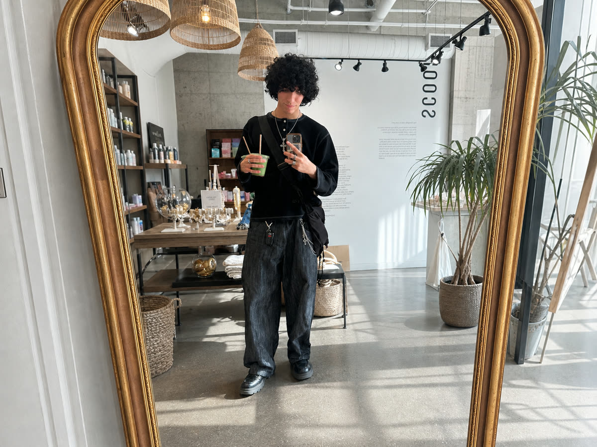 Pierre taking a mirror photo in a Toronto shop, wearing a layered black outfit and holding matcha